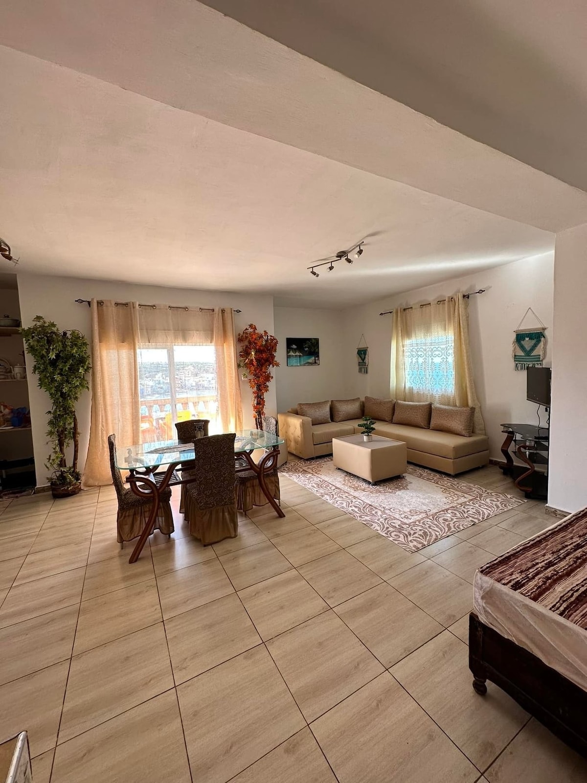 The living area features a spacious seating arrangement with light-colored sofas, complemented by a coffee table in the center. A dining table with woven chairs is positioned nearby. Natural light enters through large windows adorned with sheer curtains, illuminating the serene atmosphere.