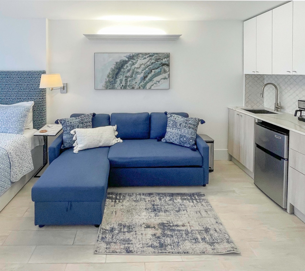 A modern studio space features a blue sectional sofa adorned with decorative pillows. A light-colored rug lies beneath the sofa, complementing the neutral tones of the flooring. Kitchenette appliances and cabinetry are neatly arranged in the background, with artwork depicting waves on the wall.
