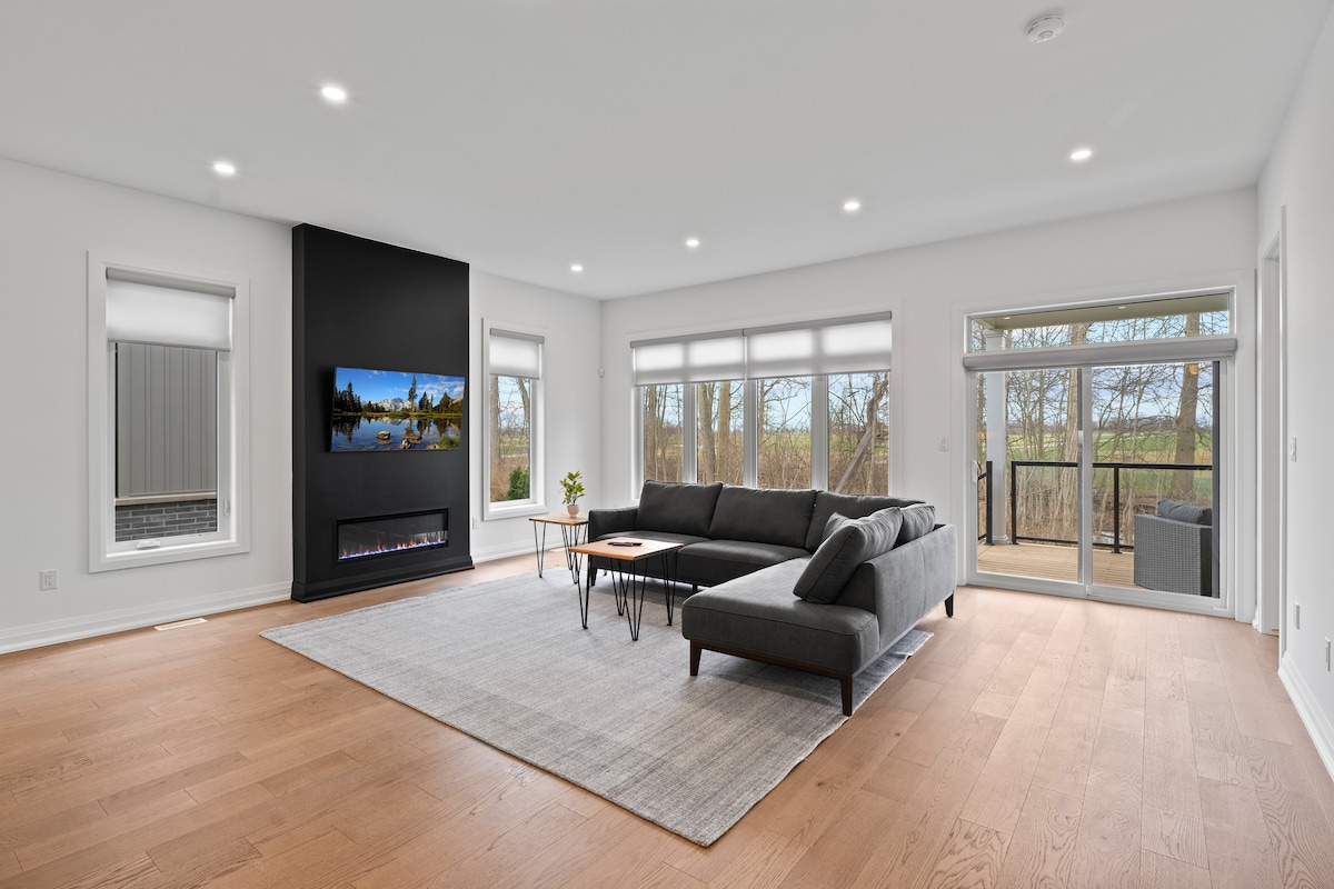 A spacious living room is featured, showcasing a large sectional sofa facing a modern fireplace and smart television. Natural light streams through expansive windows, creating a bright atmosphere. The room extends to an outdoor space with sliding glass doors that open to the serene backyard.