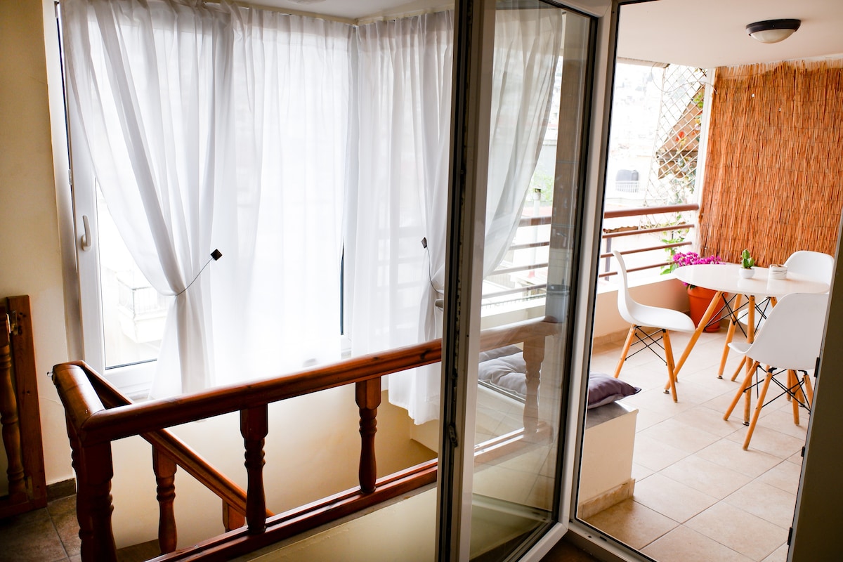 A bright balcony area is visible, featuring a wooden railing and large glass doors. Light, sheer curtains gently filter sunlight, creating a soft ambiance. A small table with chairs is arranged for outdoor dining, complemented by greenery in a planter.