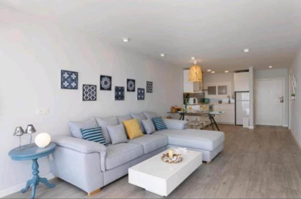 The spacious living area is furnished with a soft sectional sofa featuring decorative pillows. A white coffee table sits in front, accented by a small centerpiece. Light-colored walls are adorned with framed art, and a modern pendant light hangs above, illuminating the open kitchen in the background.