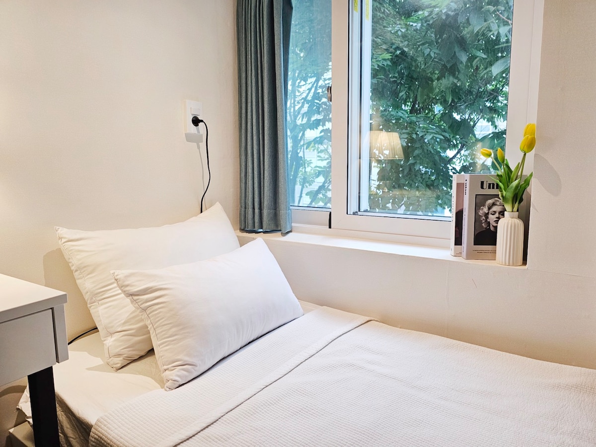 A cozy sleeping area includes a spacious bed with soft white linens and several pillows. A window with sheer curtains allows natural light to enter, while a small table displays a vase of yellow tulips and a framed photo. A calm ambience is created by the neutral walls.