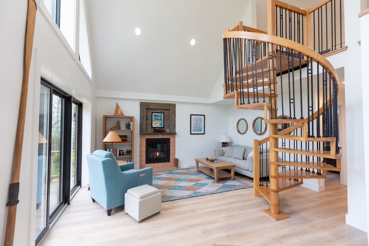 An open-plan living area features large windows that allow natural light to fill the space. A spiral staircase made of wood is a focal point, surrounded by comfortable seating and a coffee table. A vibrant area rug adds warmth, while a fireplace enhances the inviting atmosphere.