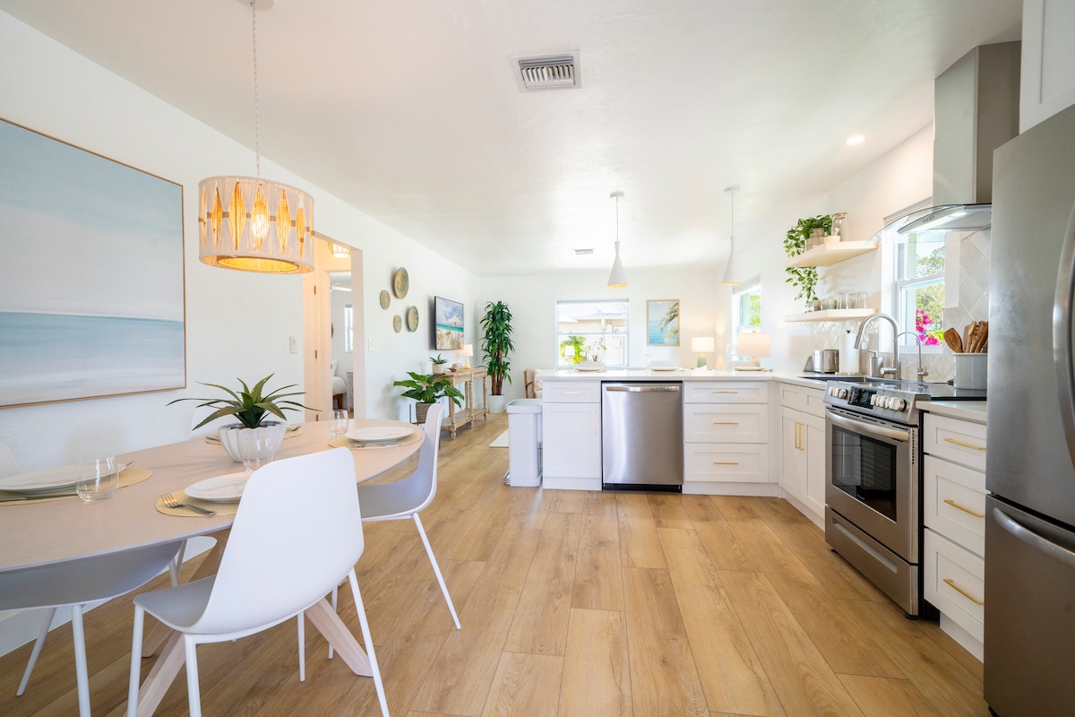 The open kitchen area showcases white cabinetry and brand-new appliances. A dining table is set for four, complemented by modern chairs. Large windows allow natural light to fill the space, while houseplants add a touch of greenery.
