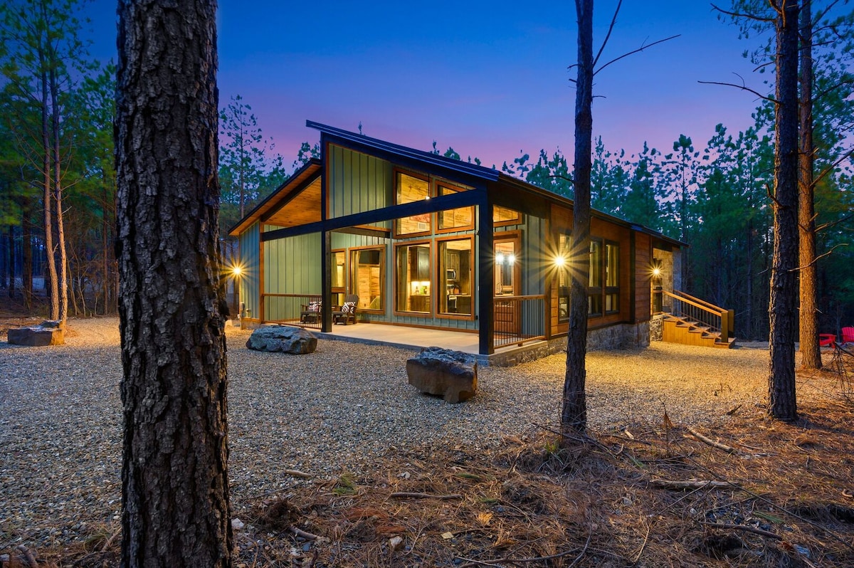 The exterior of the cabin is set amidst tall trees, featuring large windows that allow light to spill out into the surrounding landscape. A gravel pathway leads to the entrance, and stone accents are visible on the lower facade, enhancing the natural setting.