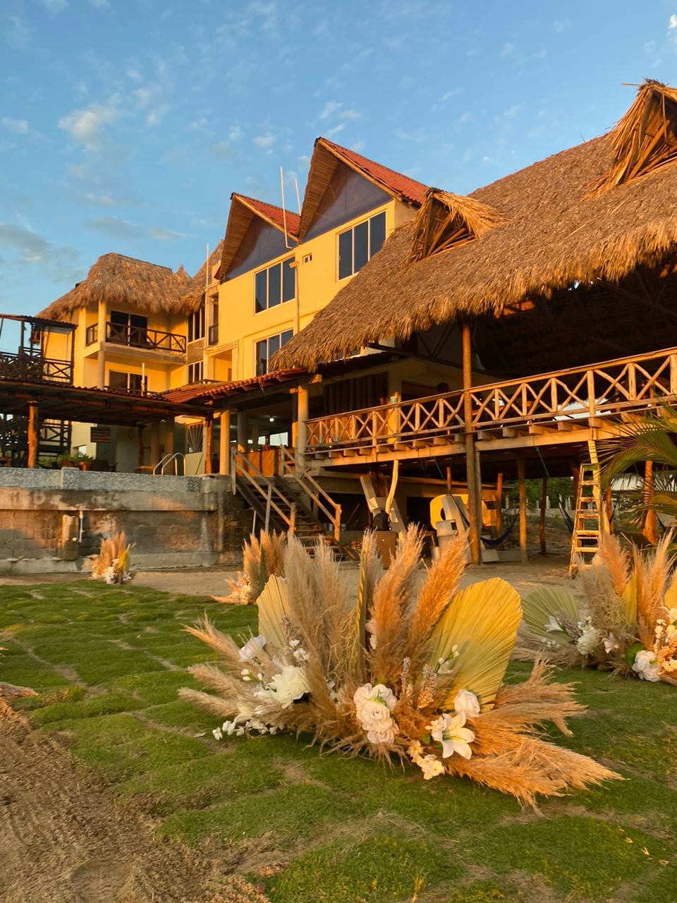 A multi-level building features a thatched roof and large windows, allowing natural light to flood the interior. Lush greenery lines the foreground, complemented by decorative floral arrangements. The warm glow of sunset casts a serene ambiance over the scene.