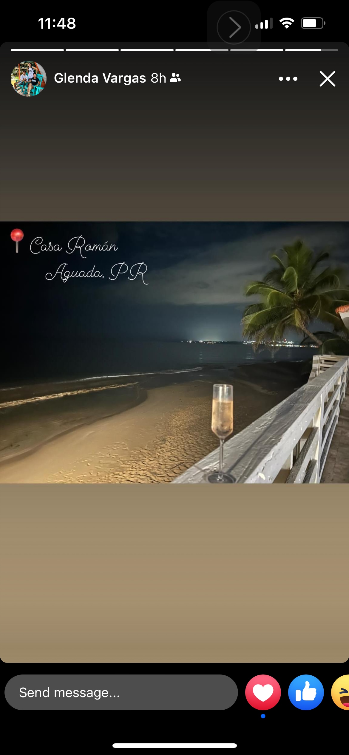 A serene evening scene is depicted with a glass of champagne on a railing. A moonlit beach extends in the background, characterized by soft waves and sandy shores. The silhouette of palm trees frames the view, and distant lights twinkle along the coastline.