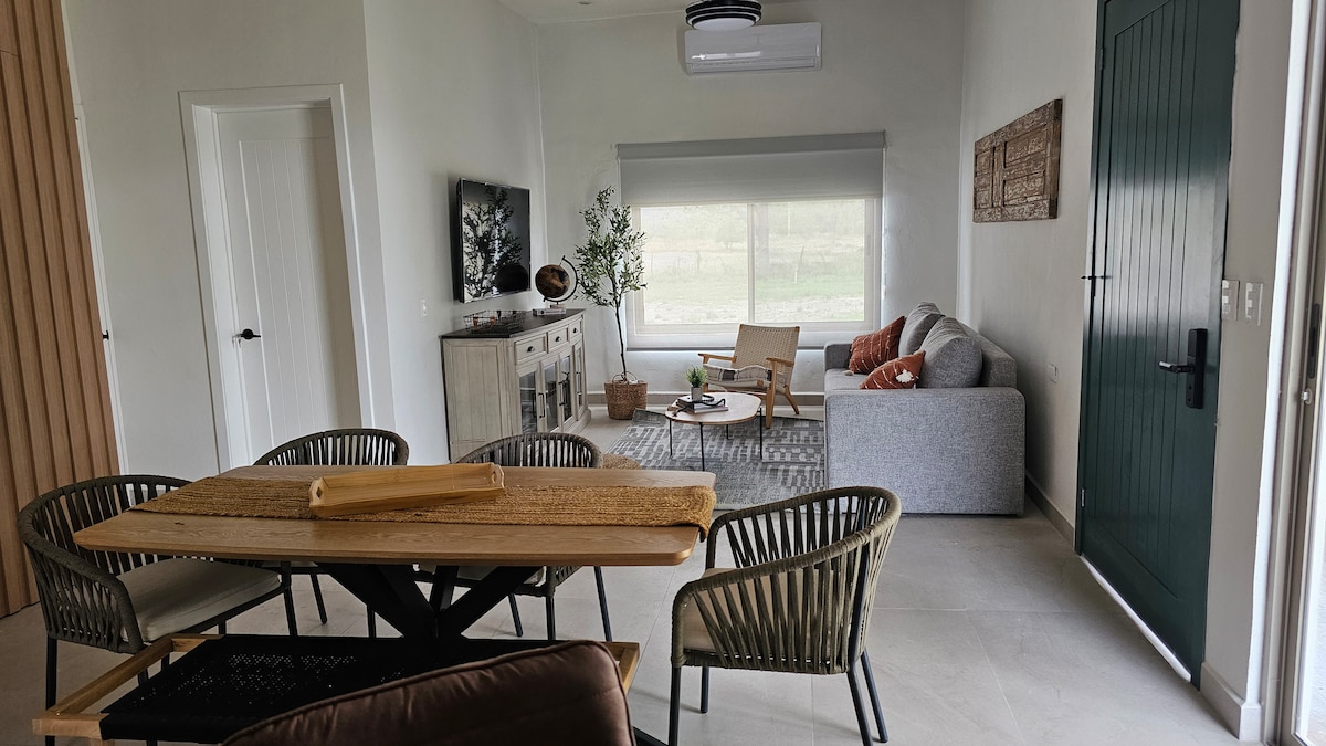 An inviting living space features a comfortable grey sofa and wooden armchairs surrounding a dining table. Natural light fills the room through a large window, offering views of the outdoors. A television is mounted on the wall, with decorative elements enhancing the atmosphere.