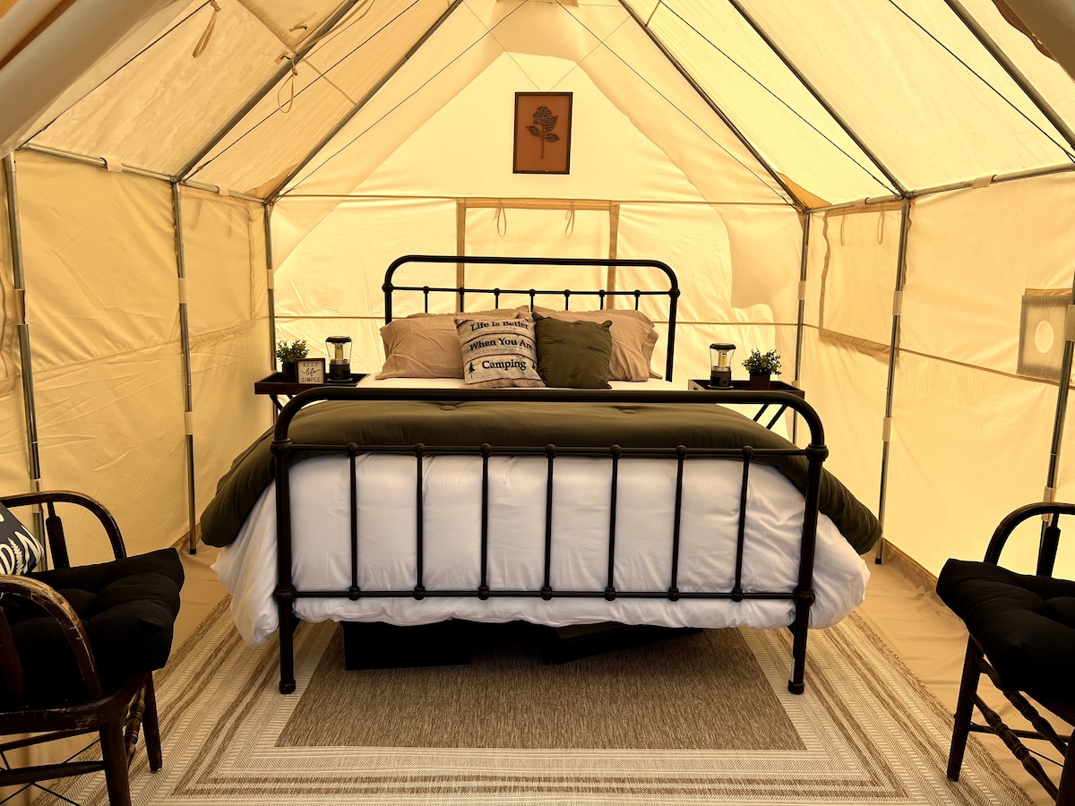 A spacious tent interior features a black metal bed framed by soft bedding in neutral tones. Two chairs are positioned beside the bed, and small plants add a touch of greenery to the space. Natural light filters through the tent's fabric walls.
