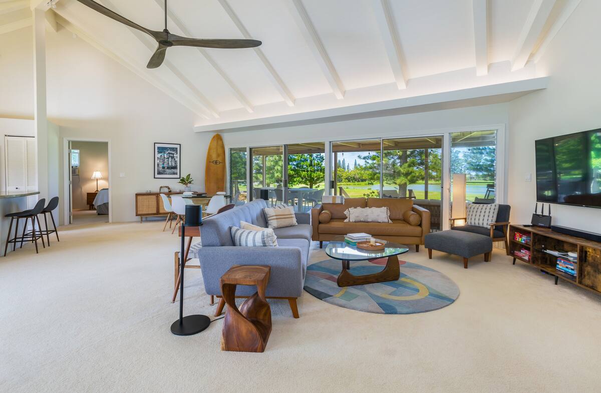 A spacious living area features high, beamed ceilings and large glass doors that open to a scenic landscape. Comfortable seating arrangements include a sectional sofa and accent chairs, surrounding a circular coffee table. A large television is positioned against the wall, accompanied by a stylish entertainment unit.