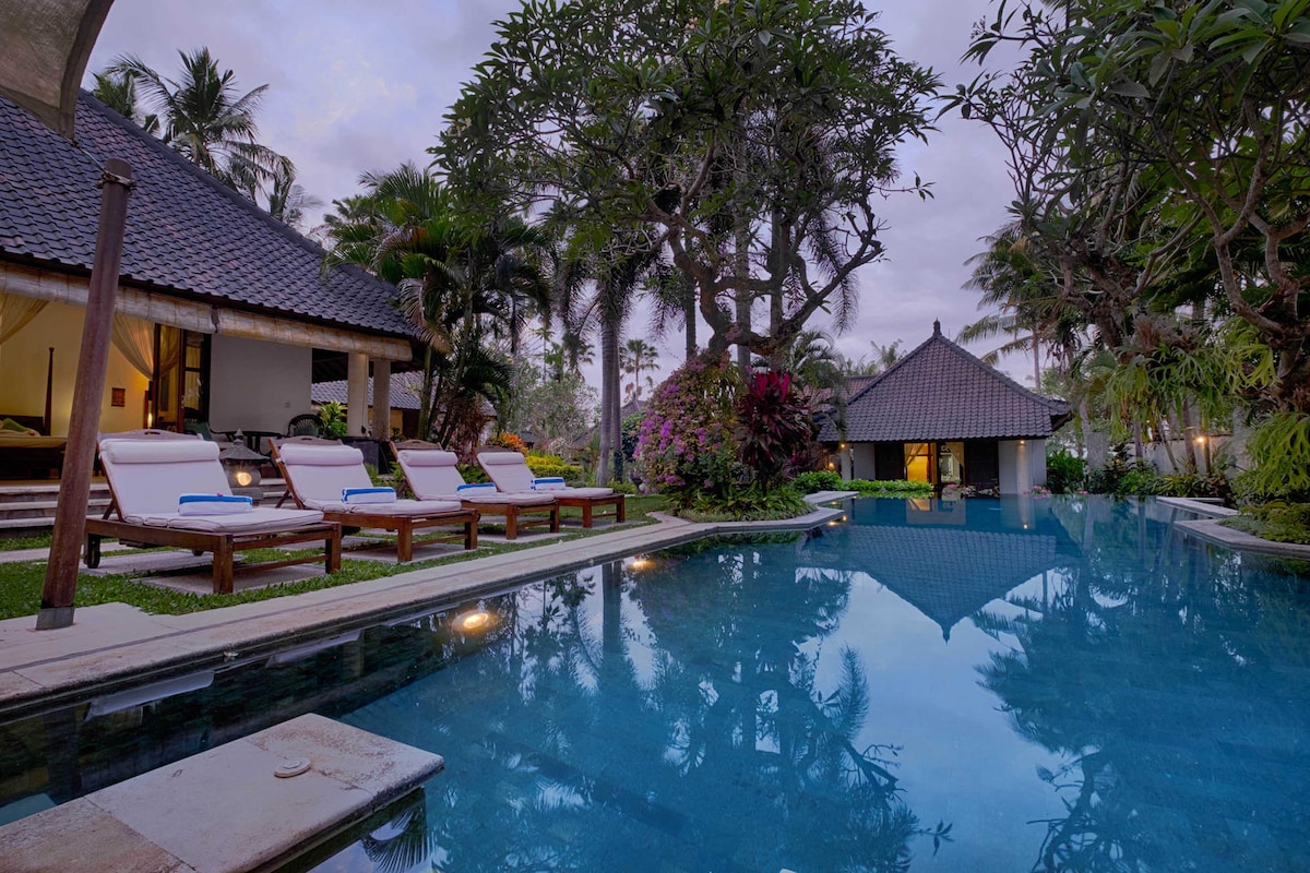 A serene outdoor area showcases a large private pool surrounded by tropical foliage. Comfortable lounge chairs are positioned beside the water. The villa's architecture features a thatched roof and is complemented by vibrant flowers and palm trees, creating a peaceful retreat.