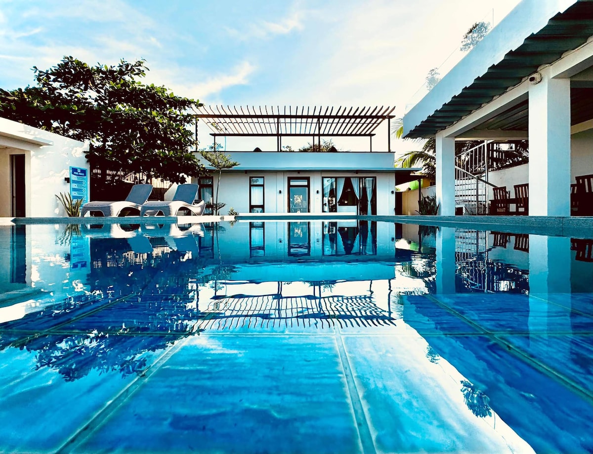 An infinity pool reflects the clear blue sky and surrounding greenery. Lounge chairs are positioned along the edge, creating a serene atmosphere. The beach house is visible in the background, framed by a pergola and tropical plants.