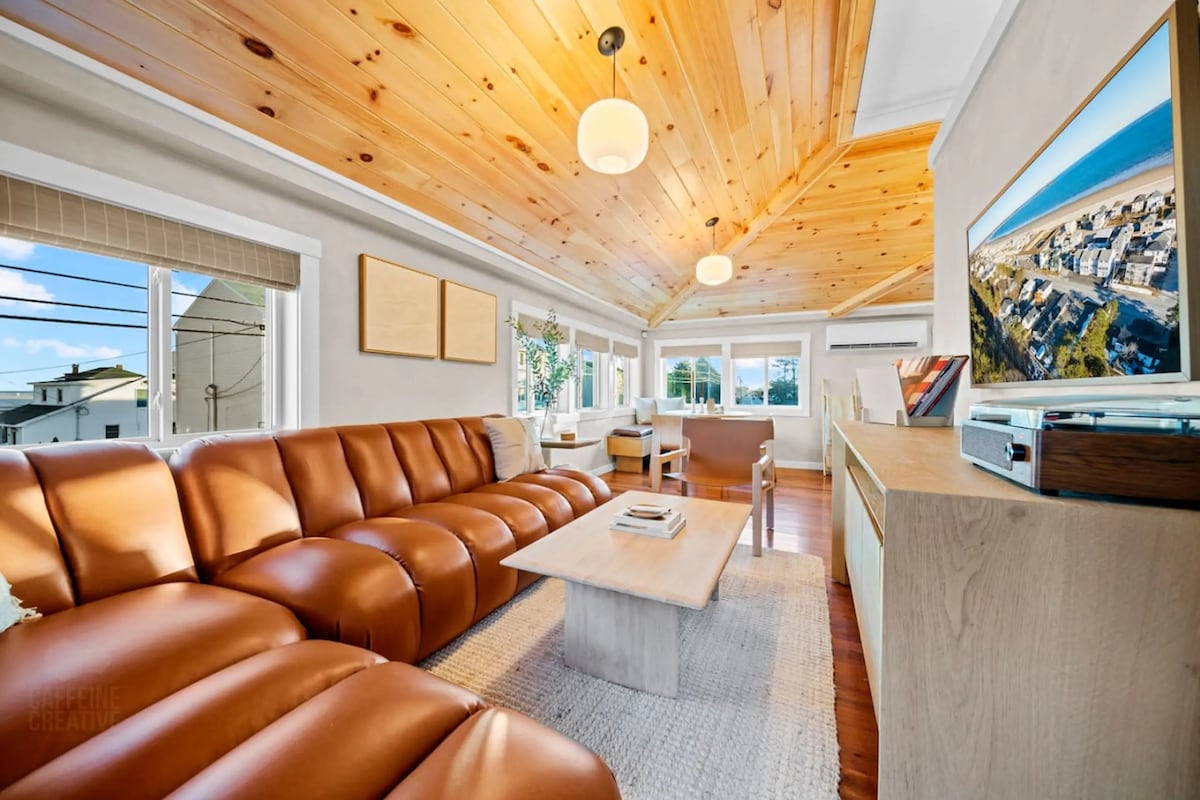 A spacious living area features a large brown leather sectional sofa arranged around a light wooden coffee table. A flat-screen television displays a coastal scene. Natural light pours in through adjacent windows, highlighting the wooden ceiling and the light-colored walls.
