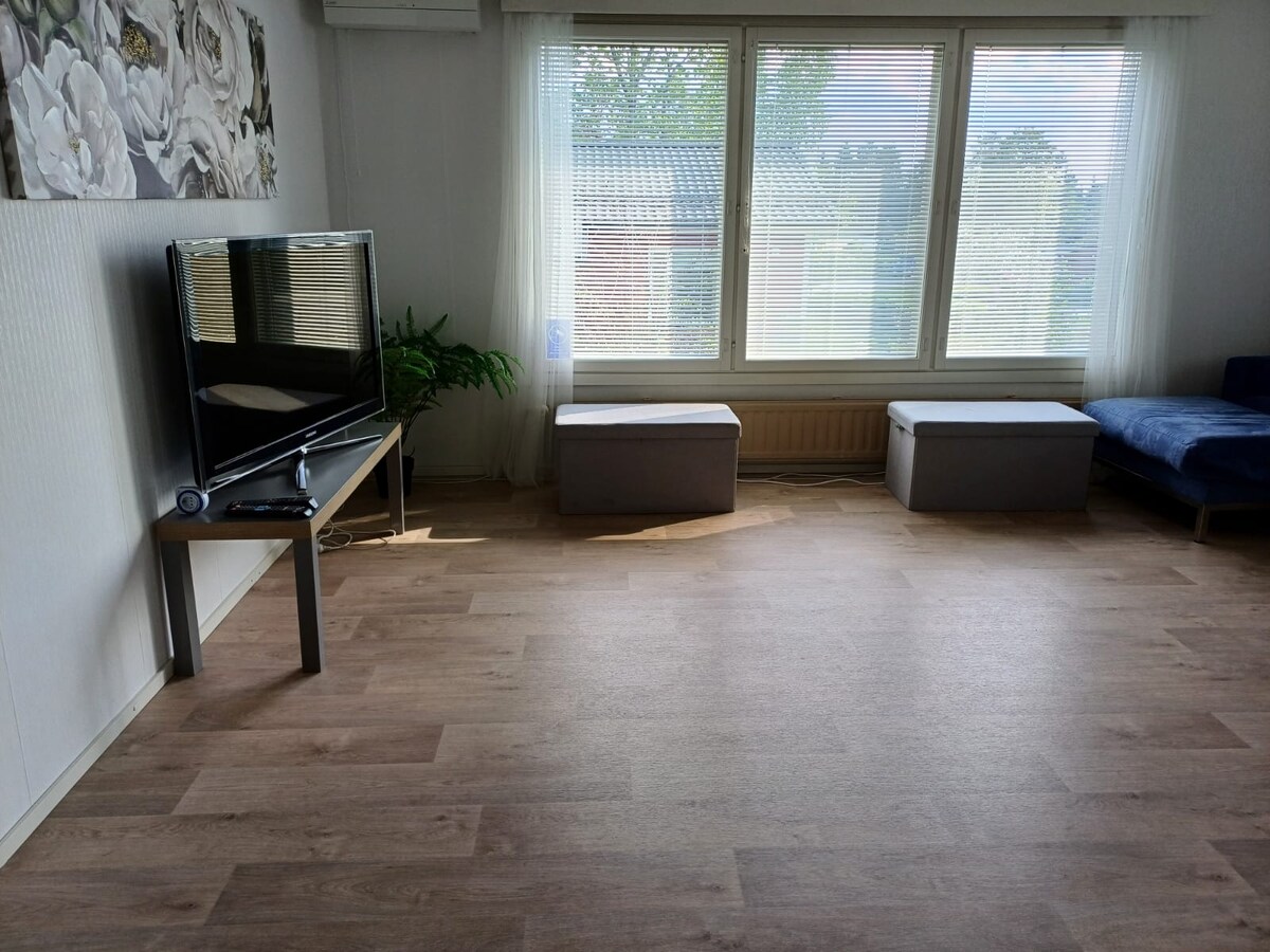 A spacious living area is presented with light-colored flooring and large windows that allow natural light to fill the space. A flat-screen television is positioned on a modern stand. Two storage benches sit opposite a comfortable sofa, enhancing the room's functionality.