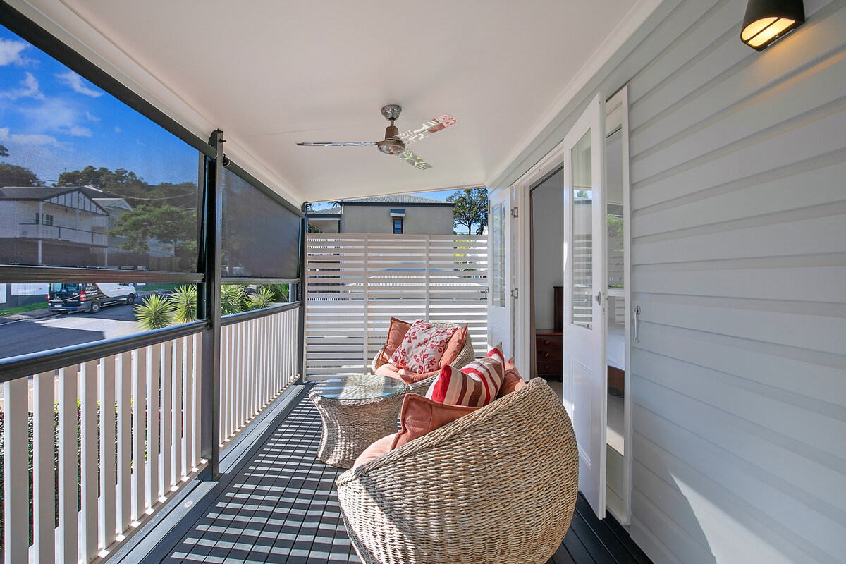 A spacious balcony features cozy seating with patterned cushions, providing a relaxed outdoor space. A ceiling fan ensures comfort, while natural light fills the area through the open doors. The balcony overlooks greenery and a charming neighborhood.