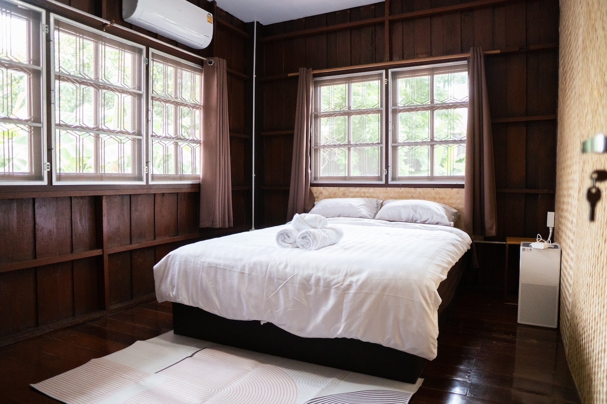 A cozy bedroom is furnished with a spacious bed topped with layered white linens and neatly arranged towels. Warm wooden walls enhance the inviting atmosphere, while natural light pours in through multiple windows, accompanied by soft, neutral curtains. A small nightstand and a refrigerator are also visible.
