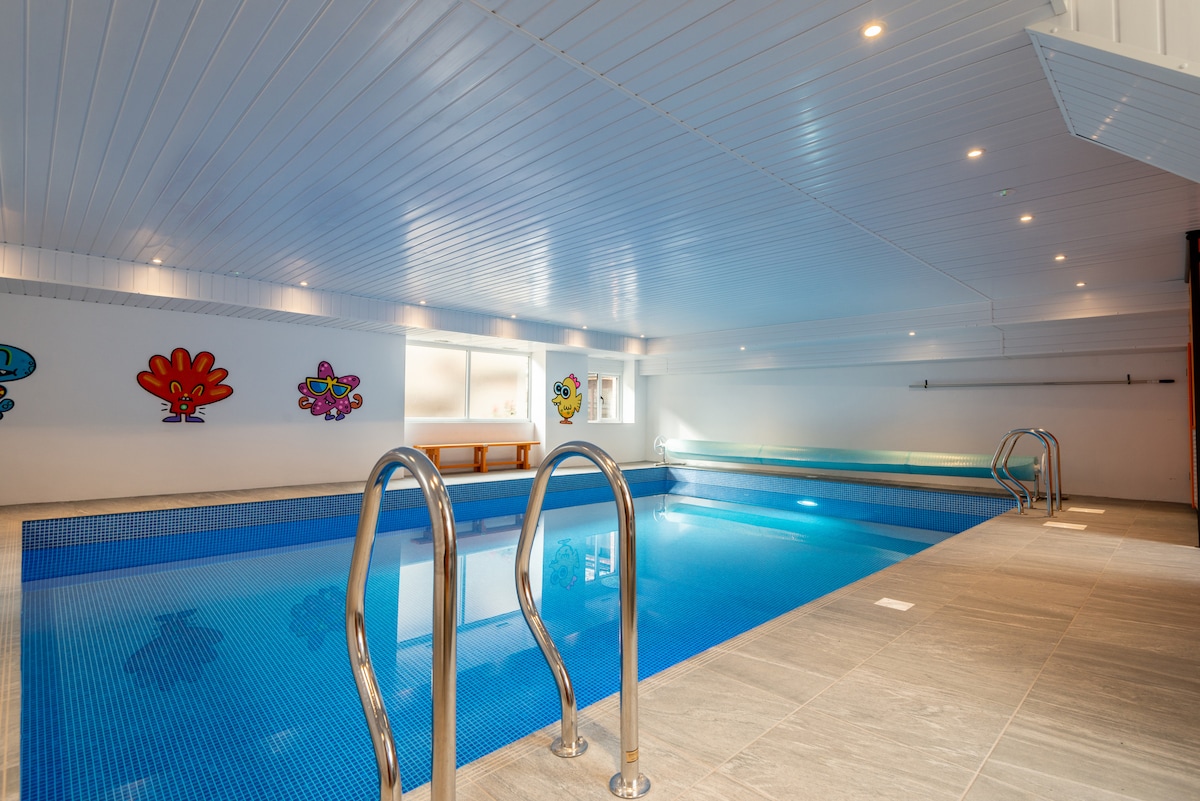 An indoor heated swimming pool is shown, featuring light blue tiles that reflect the water's surface. Decorative art pieces adorn the white walls, enhancing the modern ambiance. Soft lighting illuminates the space, and steps lead into the pool from one end.