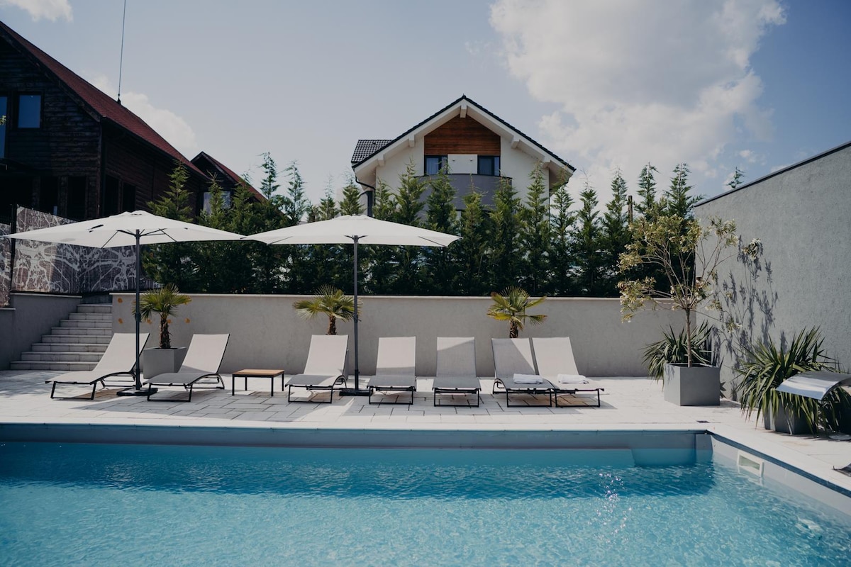 A well-maintained outdoor area features a rectangular heated pool surrounded by six lounge chairs. Two large umbrellas provide shade, and various plants are arranged around the poolside. A multi-level structure can be seen in the background, with trees offering privacy.