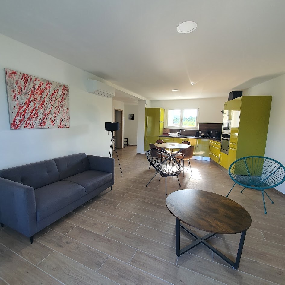 A spacious living area features modern furnishings, including a gray sofa and a round wooden coffee table. An adjacent dining space with a light-colored table and mixed seating is visible, along with green cabinetry and a fully equipped kitchen backdrop. Natural light enters through a window.