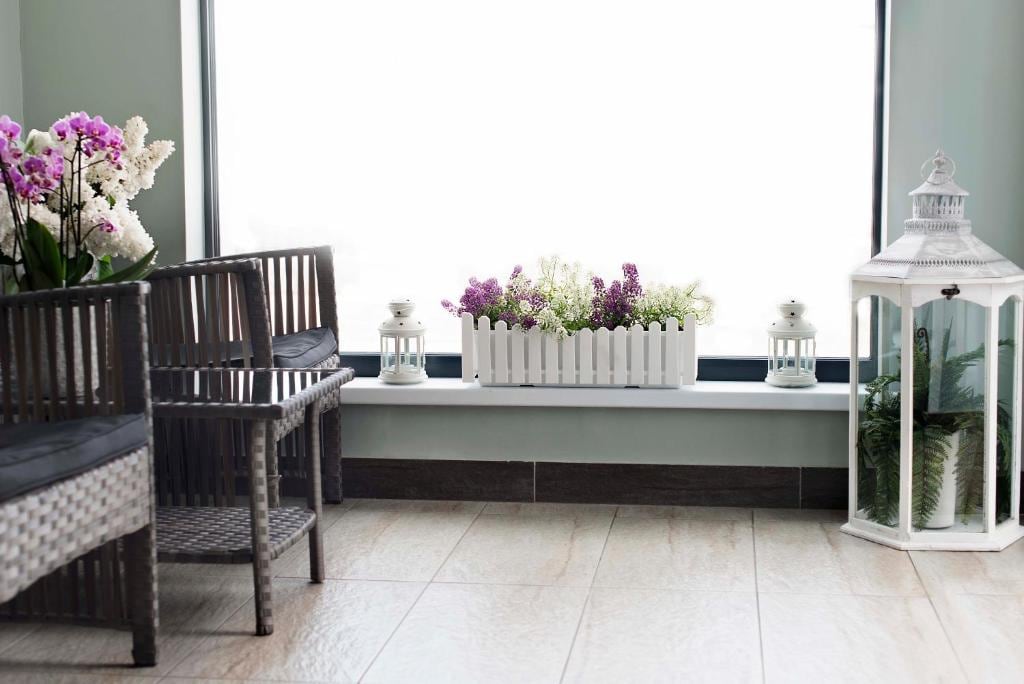 A bright indoor space features a row of elegant chairs made of dark wicker, placed beside a large window. A wooden planter filled with purple flowers adds a touch of color, accompanied by white lanterns that sit on either side of the planter.