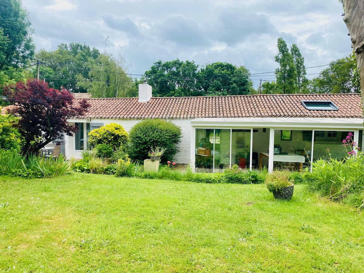 A single-story building is featured, showcasing a tiled roof and large glass windows. Lush green grass and various plants surround the structure, providing a natural setting. The outdoor area includes a landscaped garden, adding to the serene atmosphere.