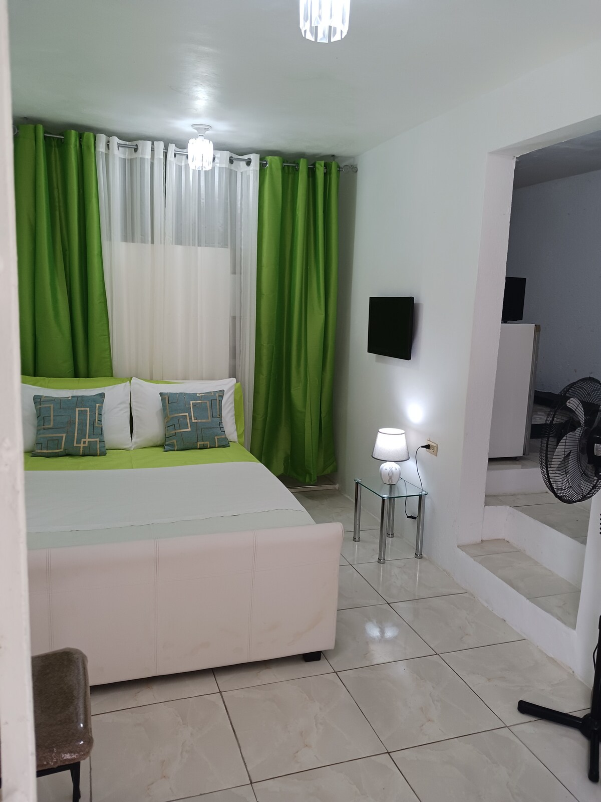A cozy room features a spacious bed adorned with decorative pillows. Green curtains frame a bright window, allowing natural light to illuminate the space. A wall-mounted Smart TV is visible, along with a small lamp on a side table and a standing fan nearby.