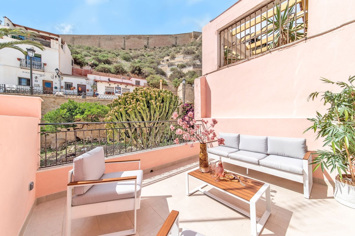 A sunlit terrace features a modern seating arrangement with light-colored cushions and a wooden coffee table. Lush green plants, including a large cactus, provide a vibrant backdrop, while the soft pink walls of the apartment enhance the space. The castle in the distance adds a historic touch.