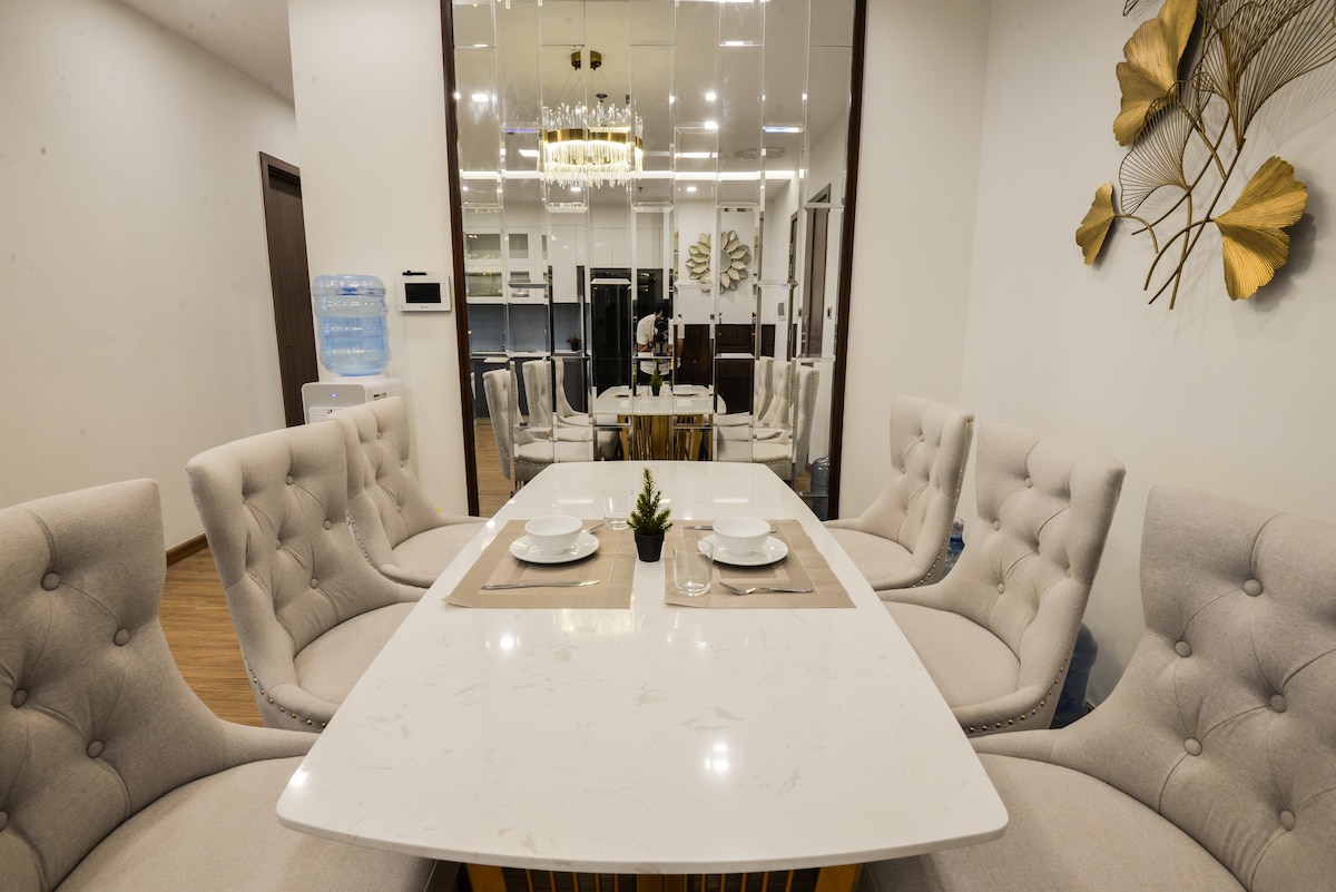 A modern dining area features a white marble table surrounded by six plush, upholstered chairs. Decorative elements include a neutral-colored table runner and simple tableware, while a mirrored wall adds depth and brightness to the space.