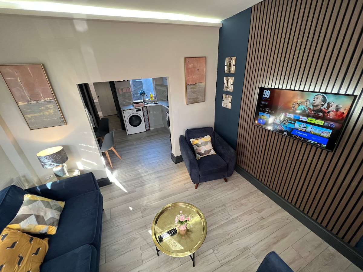 The living area features a dark blue squishy sofa positioned near a 42" smart TV displaying streaming options. A round coffee table with floral arrangement is located in the center. Behind, a doorway leads to the kitchen and laundry area, with light streaming in from above.