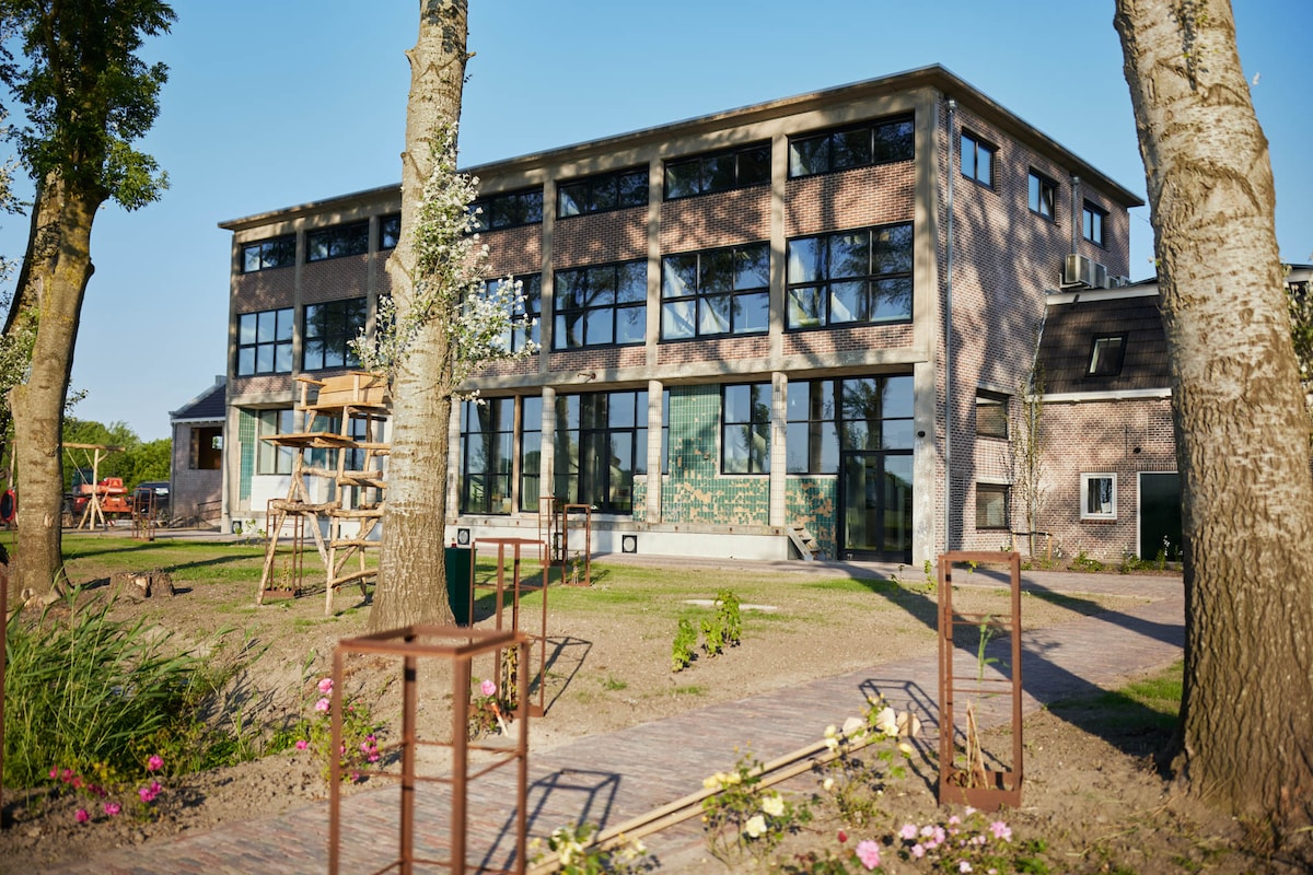 A modern, two-story building is set amidst greenery, featuring large windows that allow ample natural light. A pathway of bricks leads through a landscaped area with flowers and trees, contributing to the tranquil environment surrounding the apartment.