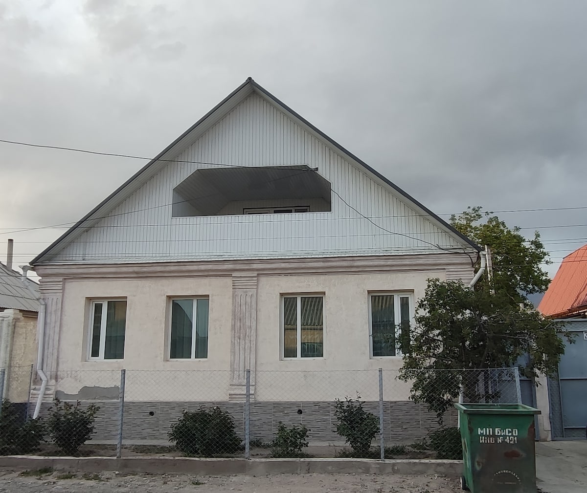 The guesthouse features a two-story structure with a light-colored exterior and a prominent balcony on the upper level. Large windows are present on both floors, and simple landscaping with shrubs is visible in front. A fence encloses the property.
