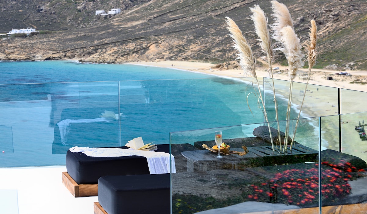 A spacious terrace features comfortable loungers overlooking a serene beach. A glass table holds a refreshing beverage and a small plate. The area is adorned with tall pampas grass, framing the stunning view of the turquoise sea and sandy shore below.