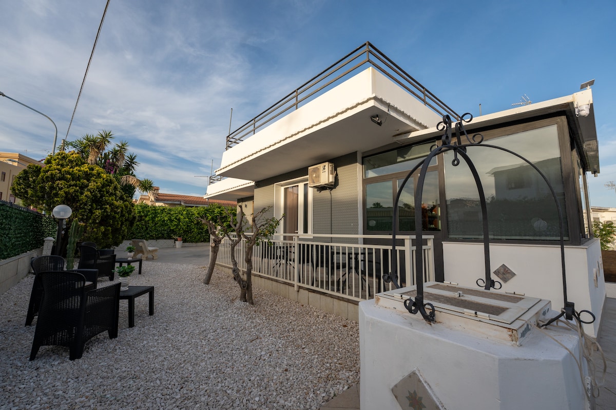 The exterior features a contemporary building surrounded by a well-maintained garden with pebbled pathways. A seating area with black chairs and a small table is visible, and a decorative well adds charm to the landscape. Lush greenery provides privacy and serenity.