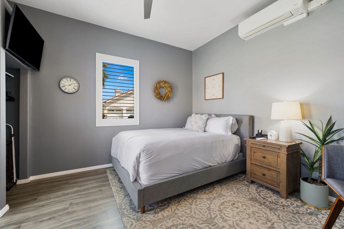 A cozy sleeping area features a queen-size bed dressed in crisp white linens. Soft gray walls provide a calm backdrop, complemented by wood furnishings, including a bedside table with a lamp. A large window invites natural light and fresh air, framed by light curtains.
