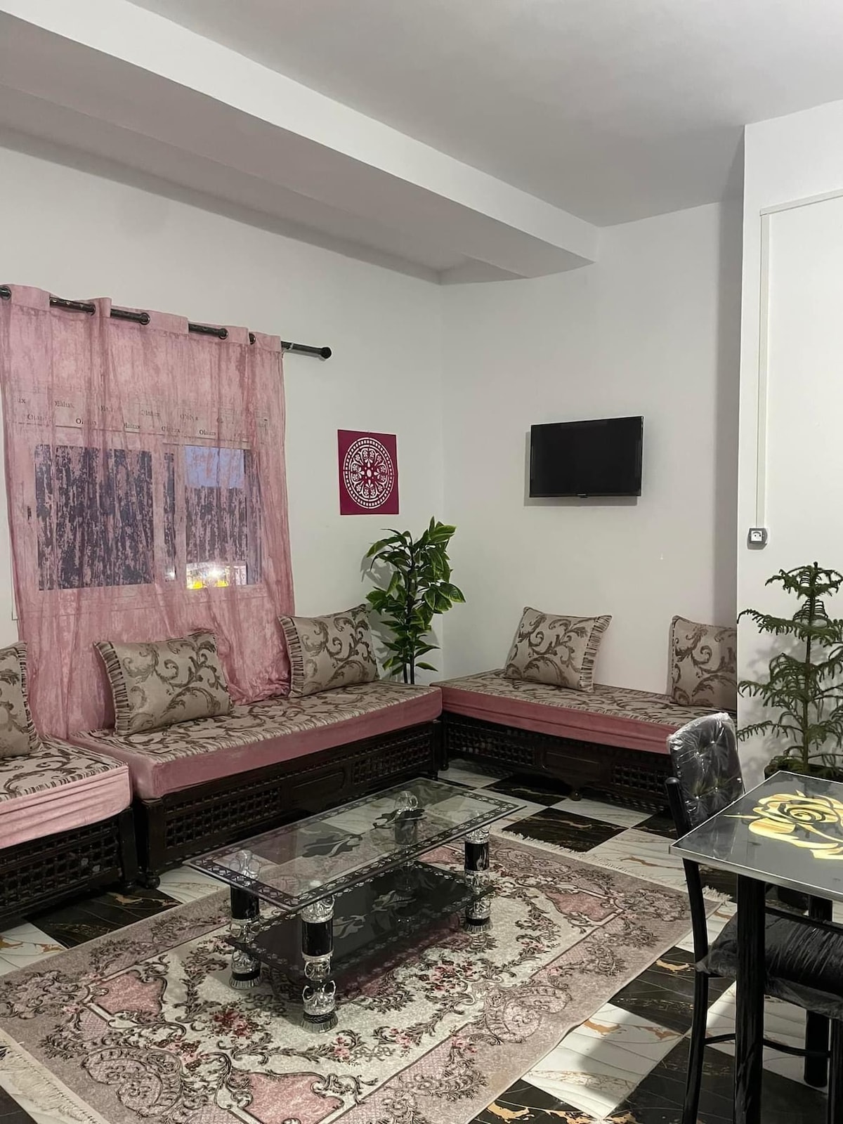 A welcoming living area is furnished with a large, L-shaped seating arrangement featuring patterned cushions. A glass coffee table sits in the center, and a small TV is mounted on the wall. Natural light enters through a window draped with light pink curtains.