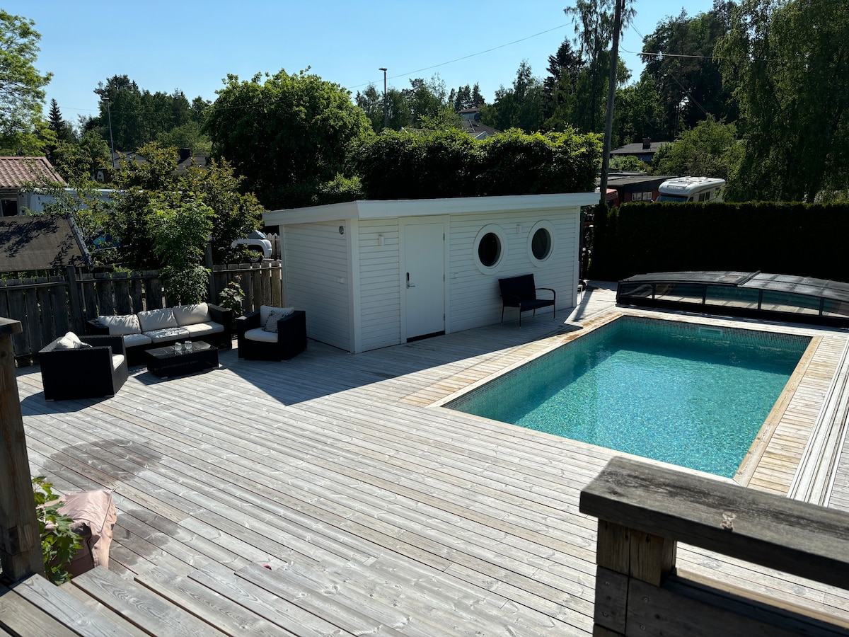 A saltwater pool is surrounded by a spacious deck made of wooden planks. A white pool house with two circular windows is visible, along with a seating area featuring dark furniture, providing a relaxed ambiance. Lush greenery borders the space, enhancing privacy.