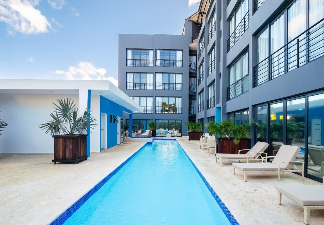 An outdoor area features a long, rectangular pool surrounded by lounge chairs and tropical plants. The building's modern facade with large windows reflects the sky, providing a lively atmosphere. Sunlight illuminates the space, enhancing the appealing ambiance of the courtyard.