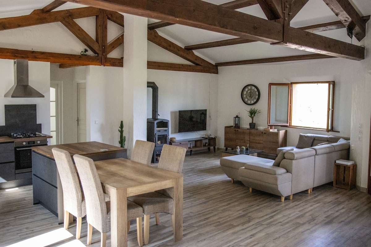 An open-concept living area features exposed wooden beams and ample natural light from large windows. A dining table with four chairs sits adjacent to a kitchen area, while a comfortable sectional sofa is positioned near a television. A rustic cabinet adds additional storage.