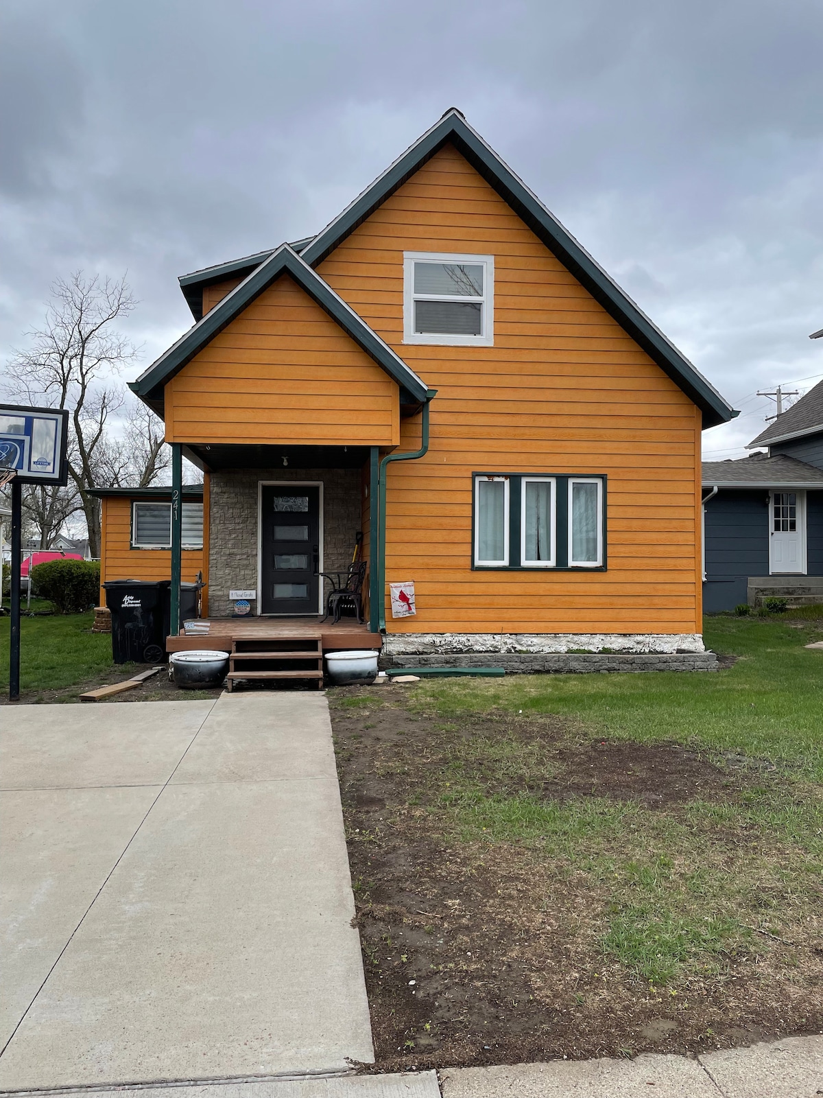 A two-story, orange wooden house features a welcoming entrance with a small porch and steps leading to the front door. Large windows allow light to enter, while a basketball hoop stands to one side. The yard is green and well-kept, with a driveway visible in the foreground.