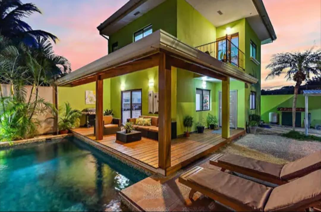 The exterior of the home showcases a vibrant green facade, enhanced by a covered patio area featuring comfortable seating. A private pool reflects the evening sky, while two loungers invite relaxation. Lush foliage surrounds the property, providing a peaceful ambiance and a connection to nature.