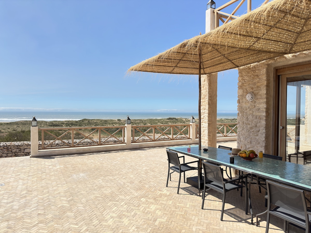 A spacious outdoor terrace features a large dining table with comfortable chairs, shaded by a natural straw awning. The setting offers a panoramic view of the ocean and surrounding landscape, enhancing the connection to nature.