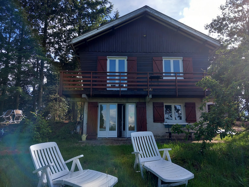 The two-story chalet features a wooden structure with a balcony adorned by railing. White lounge chairs are positioned on the grassy area, offering a view of the surrounding trees and a serene outdoor space. Large glass doors provide access to the interior.