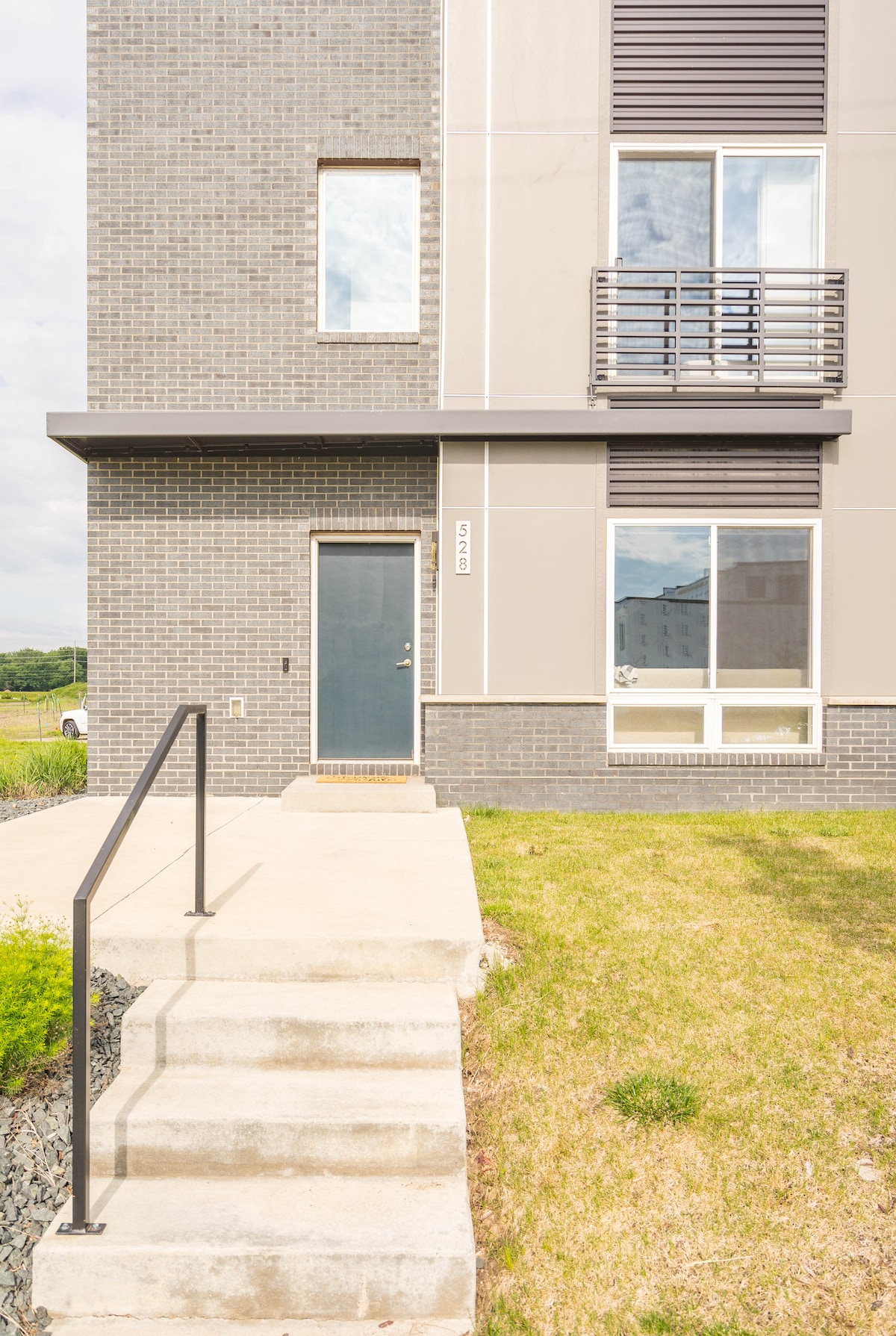 The exterior entrance of the building features a clean concrete pathway leading up to a solid door with a small window above. A set of steps, accompanied by a handrail, provides access, while modern gray brick complements the overall architecture.
