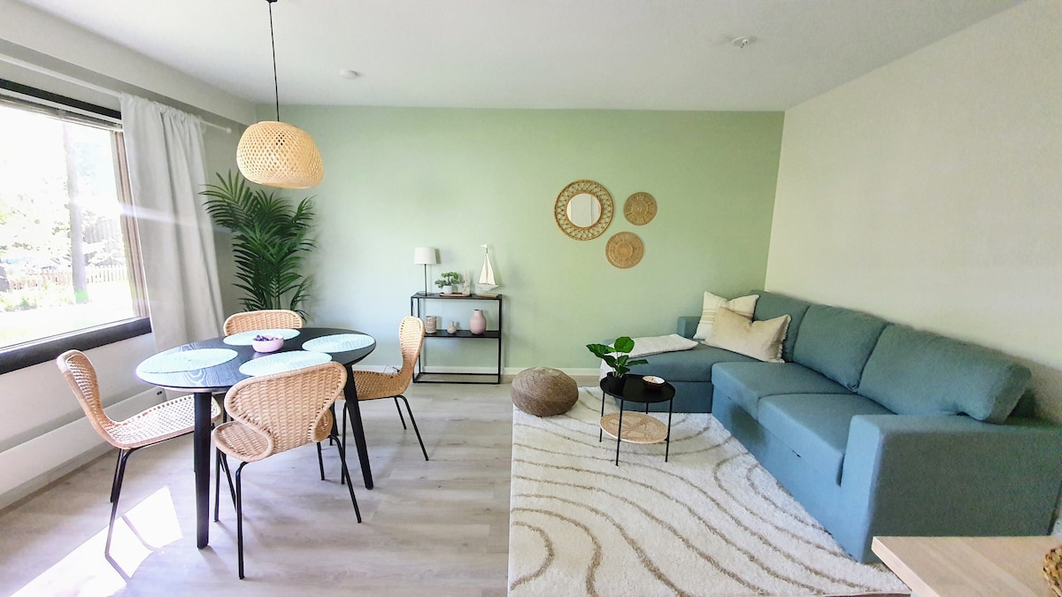 A living area is presented with a teal sofa and a round dining table surrounded by woven chairs. Natural light enters through a large window, highlighting a decorative plant. Textured wall art and a soft area rug complement the space's inviting design.