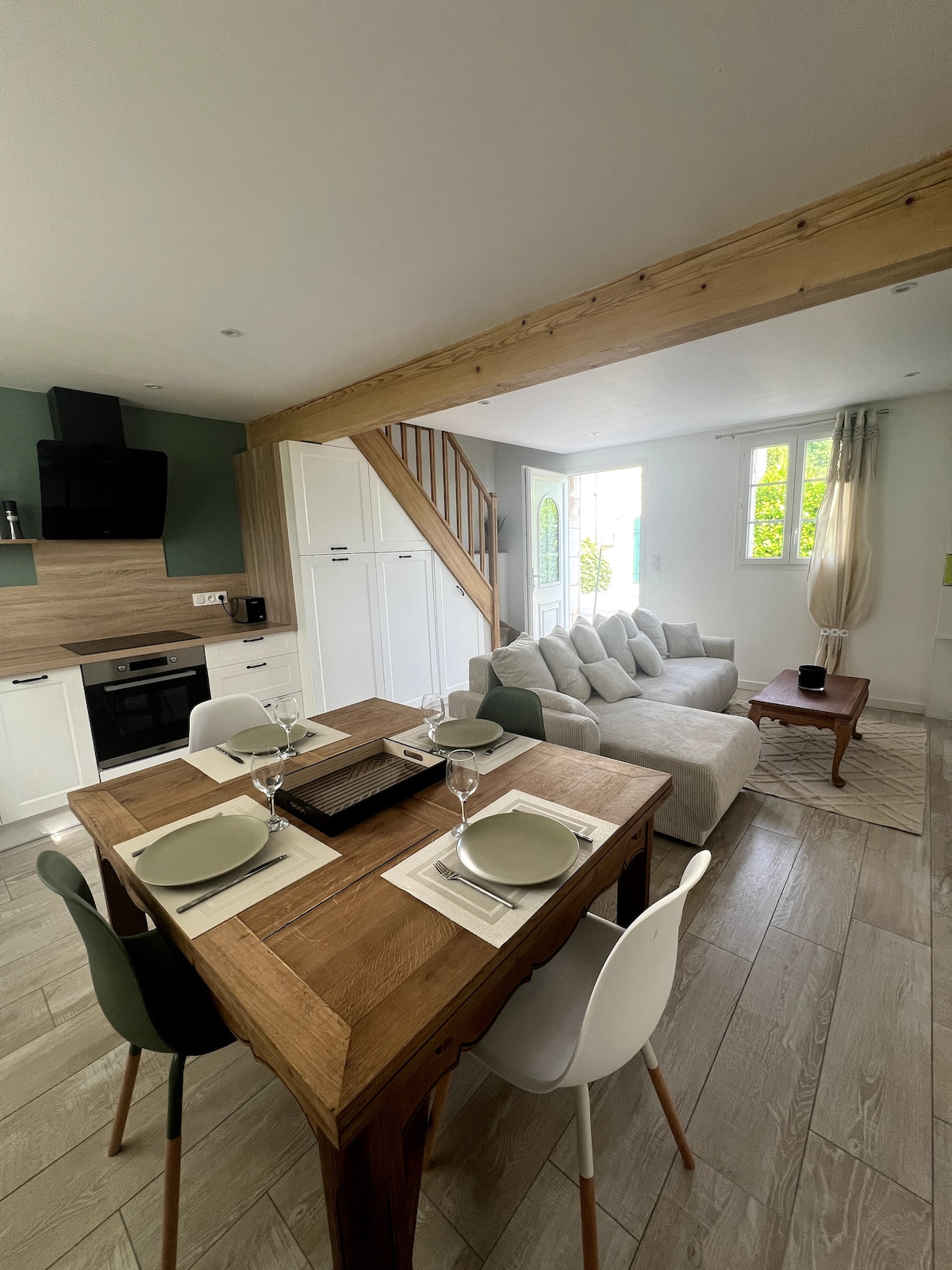 The living area features a light-filled space with a comfortable sofa and a wooden dining table set for four. The kitchen area is equipped with modern appliances. Large windows allow natural light to illuminate the room, complemented by neutral-colored decor.