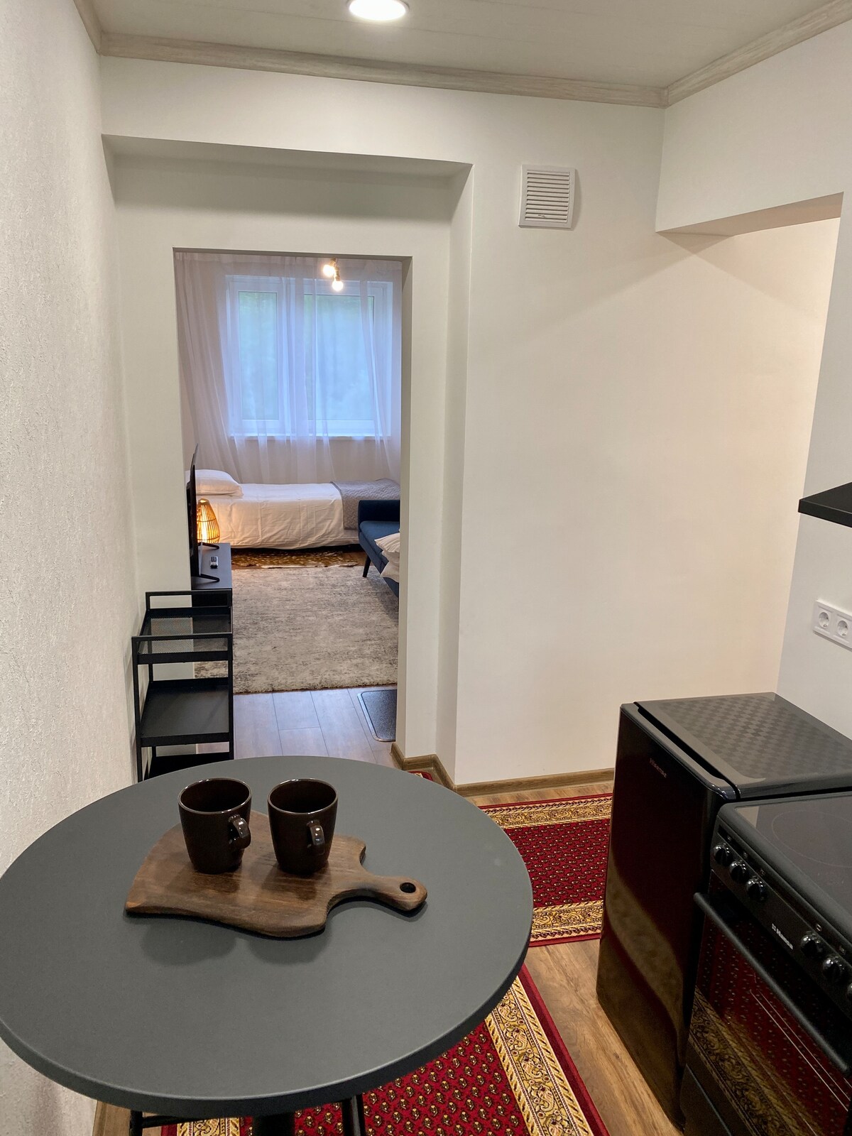 A compact kitchen area is visible, featuring dark cabinetry and a stovetop. A round table holds two coffee cups on a wooden serving board. Soft lighting enhances the neutral walls, leading into a cozy living space that includes a bed and a plush area rug.