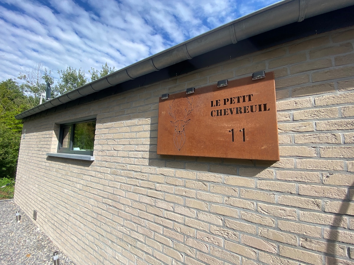 The exterior of a modern building is depicted, featuring a stone facade and a metal sign reading 'Le Petit Chevreuil' accompanied by a deer illustration. Natural surroundings are visible, enhancing the peaceful environment.