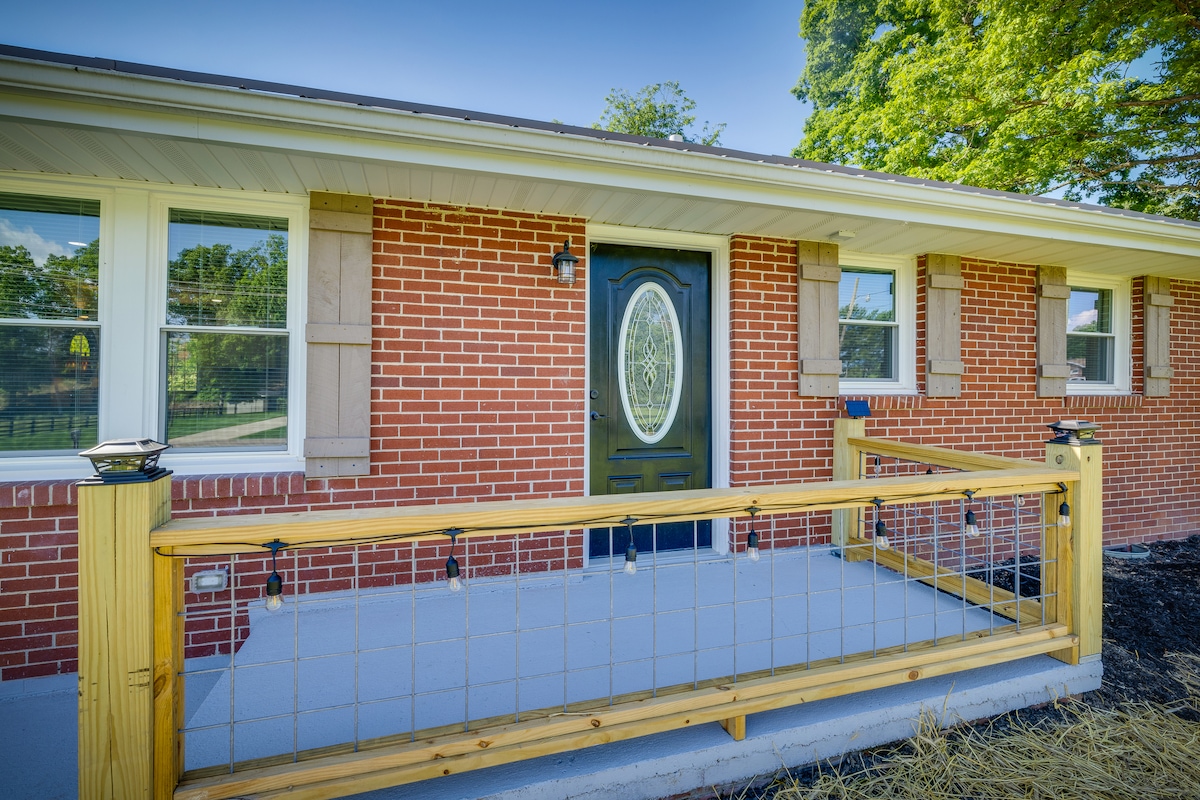 A welcoming front porch is displayed, featuring a sturdy wooden railing with decorative lighting. The brick exterior of the house is complemented by large windows and a prominent oval door, enhancing the inviting entrance.