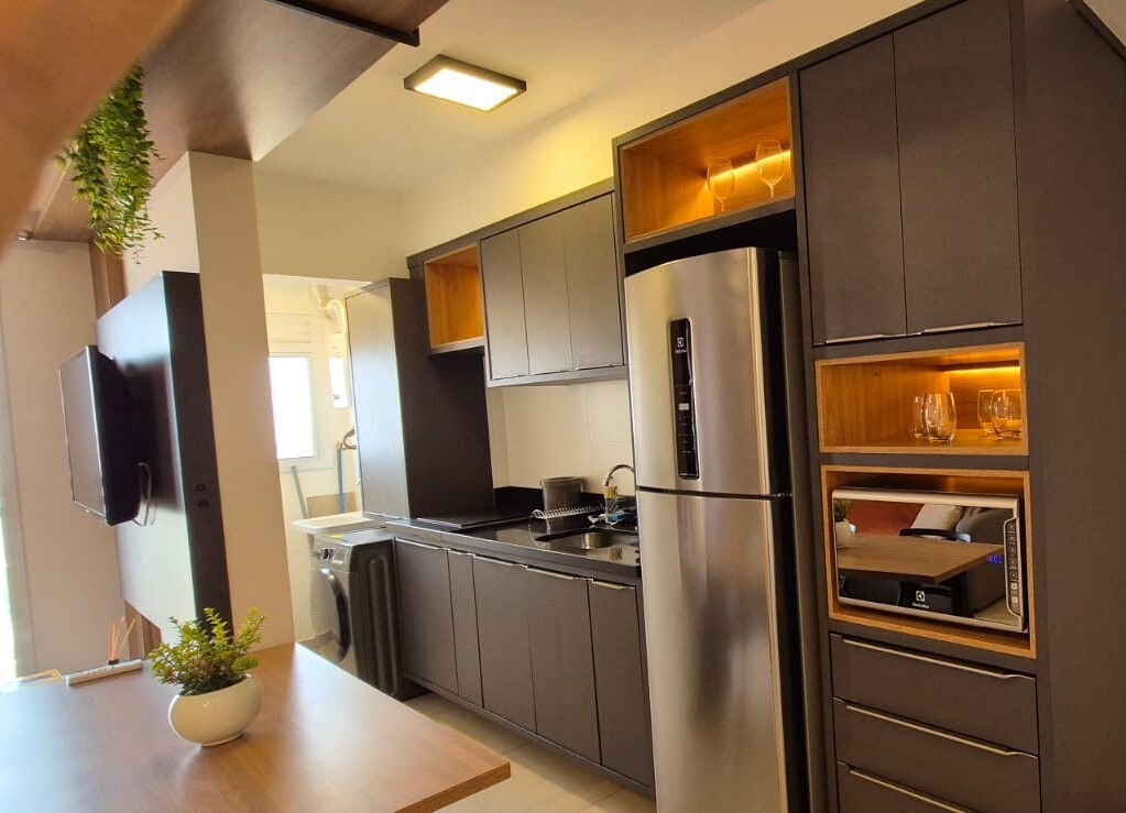 The kitchen features modern cabinetry with contrasting warm wood accents. A stainless steel refrigerator and sleek appliances are visible, alongside a black countertop providing ample workspace. Natural light filters in through a nearby window, enhancing the efficient layout of the space.