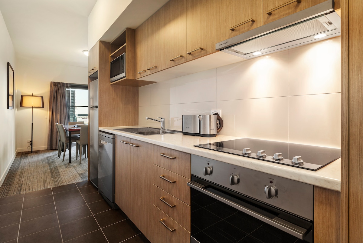 A fully equipped kitchen is visible, featuring wooden cabinetry, a stovetop, and an oven. A microwave and full-size fridge are included. The kitchen counter is complemented by a sink and modern appliances. To the rear, a dining area with grey chairs and a table can be seen.